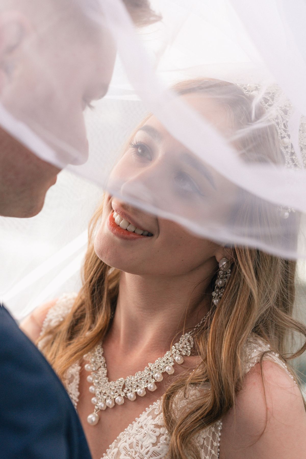 A bride smiling under her veil as the groom leans in