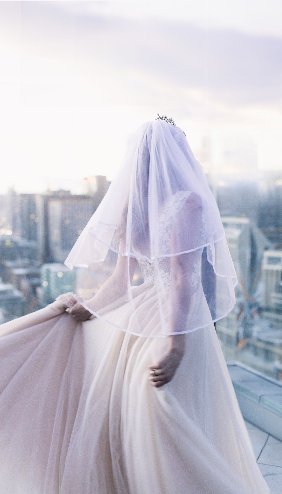 A bride from behind, holding her gown out, her veil lit by the evening sky